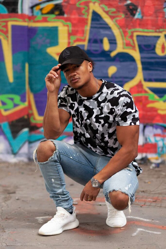 Fashion image of a man crouching on one knee wearing an urban camo t-shirt, ripped jeans, white sneakers and a black baseball cap. Brightly-colored graffiti in the background.